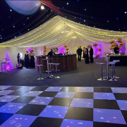 Event space with a dance floor, bar, and people gathered under a marquee decorated with white drapes and golden balloons for a corporate gala by Melody Corporation.