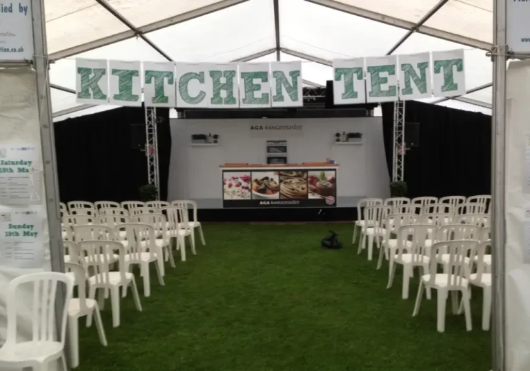 White chairs arranged facing a stage with a screen under a banner reading "kitchen tent" in a large facility tent by Melody Corporation at an event.