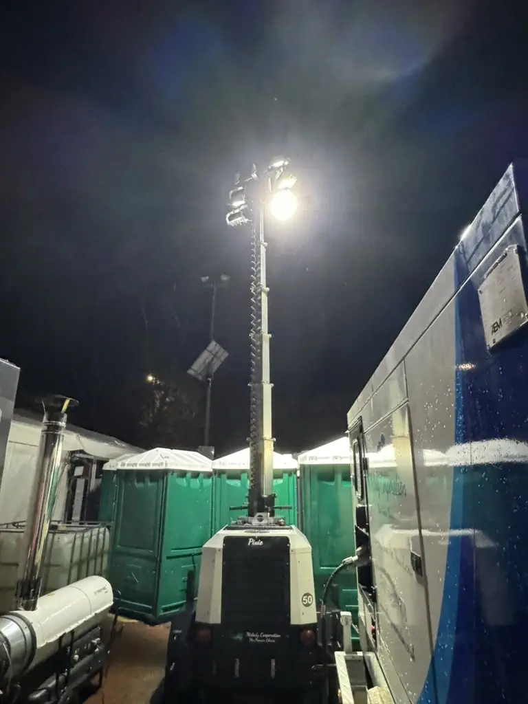 A portable lighting tower by Melody Corporation illuminates a construction site at night, surrounded by equipment and temporary facilities.
