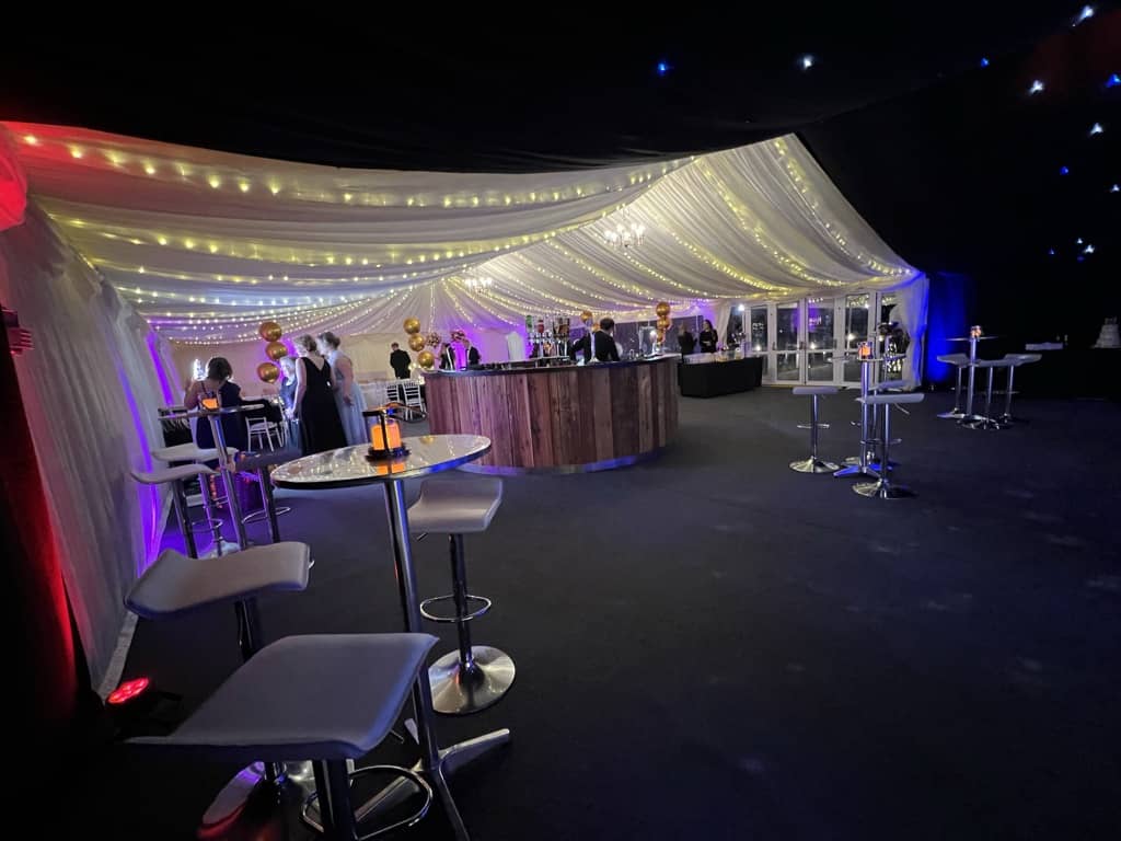 An elegant event space with a curved bar, draped white ceiling, mood lighting, and scattered high tables, occupied by guests interacting and enjoying drinks under a luxury marquee.