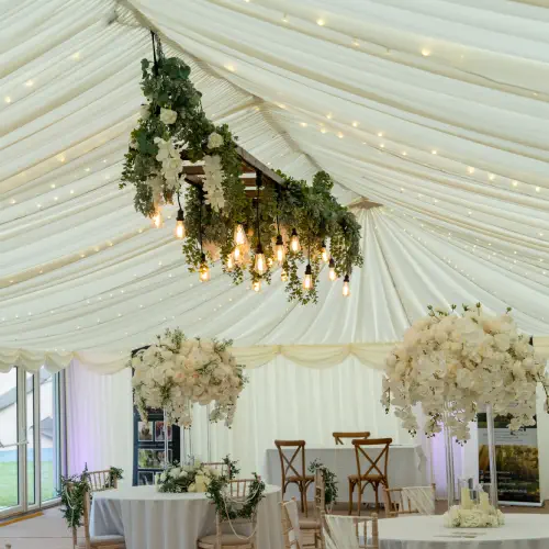 Elegant wedding venue interior with floral decorations hanging from the ceiling, white drapery, lit candles, and wooden chairs supplied by Melody Corporation.