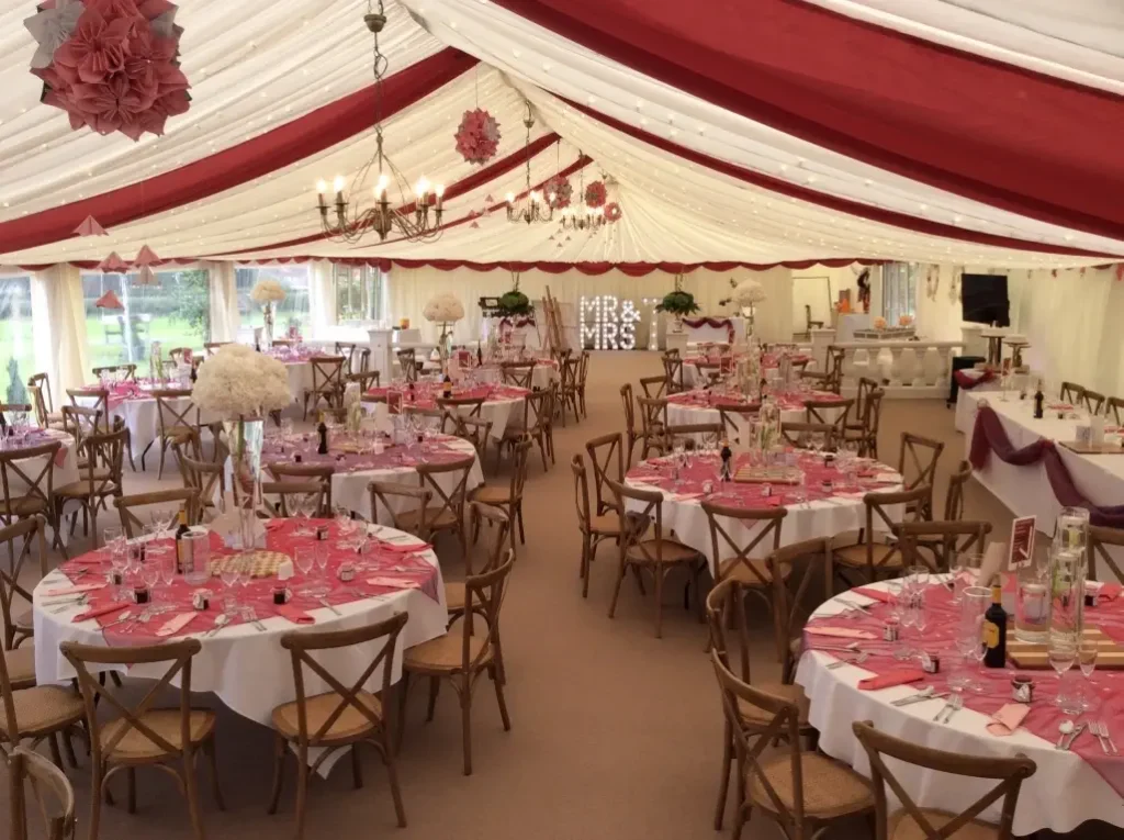Elegant outdoor wedding marquee with red and white drapery, round tables set with pink tablecloths and floral centerpieces, and a "mr & mrs" sign in the background.