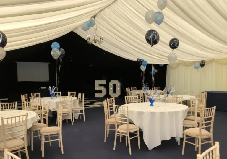 Elegant indoor party marquee by Melody Corporation with white draped ceiling, round tables with white tablecloths, and blue and black balloons for a 50th celebration.