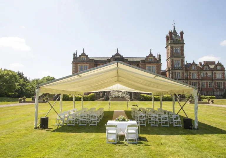 A large outdoor wedding setup with chairs and a covered stage on a lawn, with a grand historic manor house in the background under a clear blue sky provided by Melody Corporation.