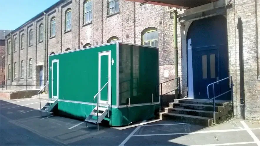 A mobile green restroom unit is placed outside a brick building adorned with multiple windows and a flight of concrete stairs, seamlessly integrating into the scene like an essential accessory at a grand marquee event.