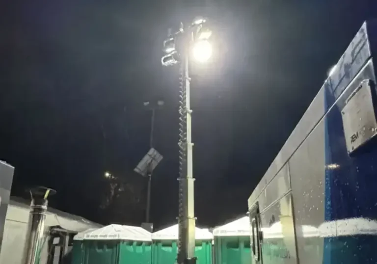 A tall, illuminated light mast at night, surrounded by portable restroom units and outdoor tents. Marquee accessories sparkle in the rain visible on the surfaces.