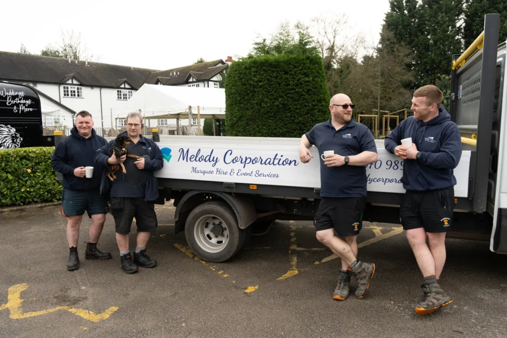 Four men in work attire stand by a Melody Corporation truck, holding mugs. One man, who is about as cheerful as can be, cradles a small dog. The truck advertises "Marquee Hire & Event Services.